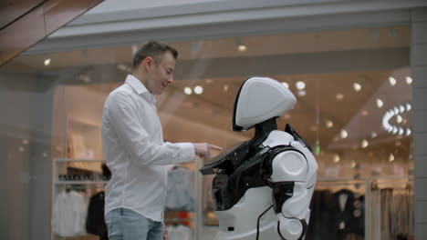 A-man-stands-with-a-robot-bot-and-asks-him-questions-and-asks-for-help-by-clicking-on-the-screen-on-the-robot-body.-Human-robot-interaction-in-the-modern-world